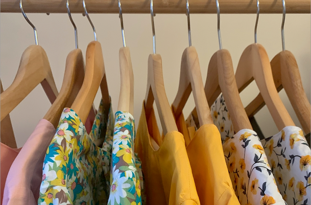 a close up of 8 dresses hanging on wooden coat hangers in different cotton fabrics, pink, blue flora, yellow and white with yellow flowers